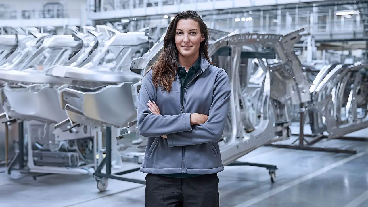 Female factory worker stands with arms crossed in an automotive assembly plant, with unfinished car frames on a rolling rack behind her, conveying a manufacturing context.